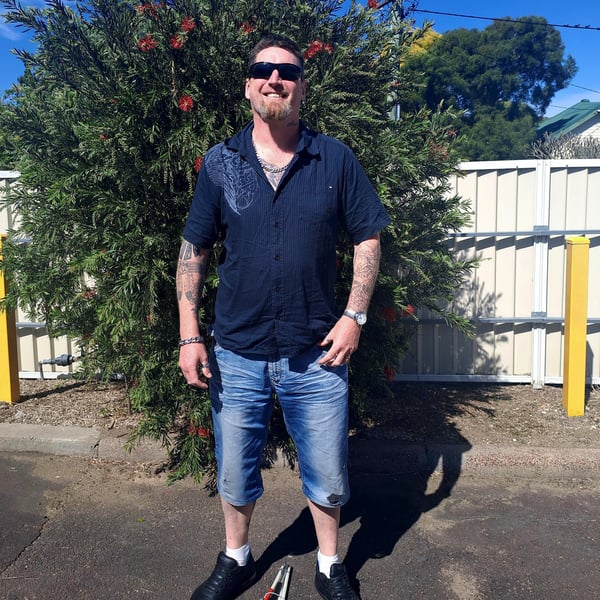 Ian has short dark hair, a goatee and tattoos on his arms. He is standing in front of a bush, smiling confidently. He is wearing ark sunglasses, navy short sleeve shirt and denim shorts. Between his feet on the ground is a folded white cane.