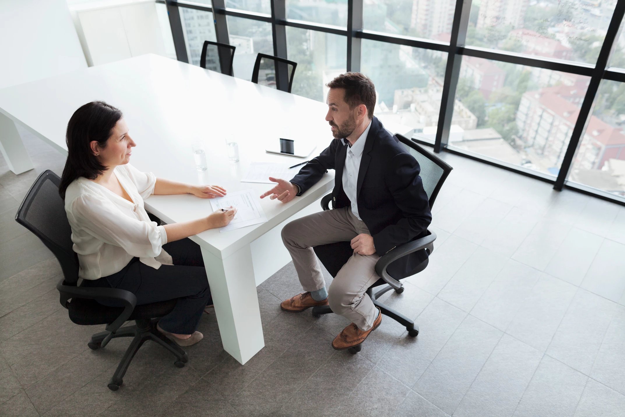 A man and a women talking. They are sitting at a large white table in front of a large window