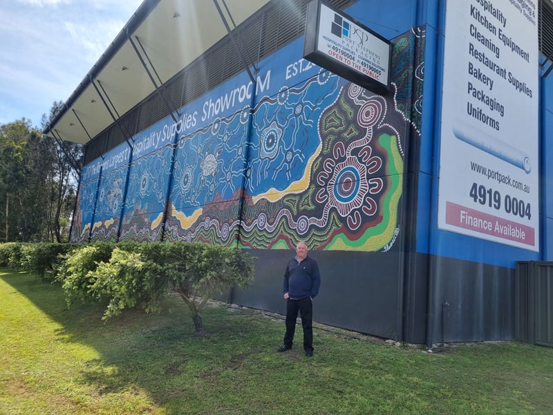 Rob is standing in front of the external wall of a warehouse, an Indigenous artwork is painted on it below the words PSP Hospitality Supplies Showroom