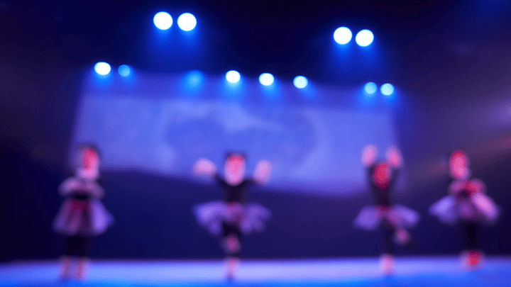 Four kids dressed in black ballerina tutus perform on stage under blue spotlights. the image is blurry and out of focus.