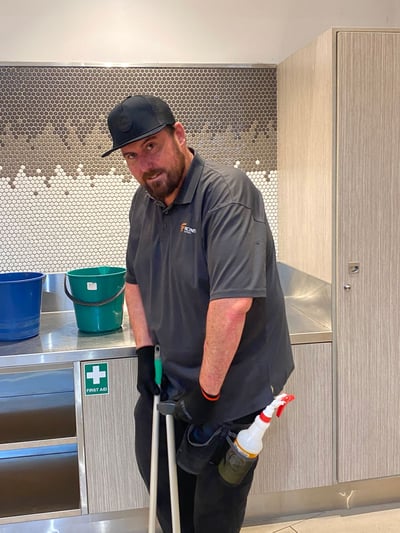 Chris is 37, he has a short beard, a black cap and is wearing a dark polo shirt, gloves and a spray bottle attached to his belt. He is holding a short broom and is standing in a kitchen area.
