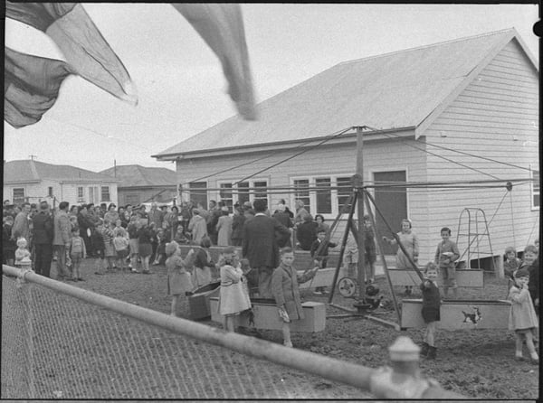 An old black and white image of people and children gathered around a white weatherboard building. The opening ceremony of Delando school in 1954. The children are playing on a clothes line transformed into a carousel.