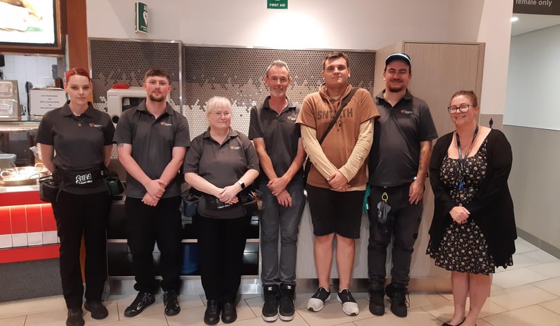 A group of people pose in the food court area of a shopping centre. They are mostly wearing grey polo shirt work uniforms.  They are cleaners. Some of them have utility belts with gloves and tools attached.