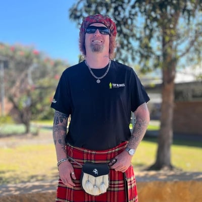 Ian is a 40 year old man with a goatee, and tattoos on his arms. He is wearing a black t-shirt with the 'Top Blokes' logo, black sunglasses, a red tartan Scottish kilt and red tartan beret hat. Ian is standing in front of a grassed area which has some trees, smiling.