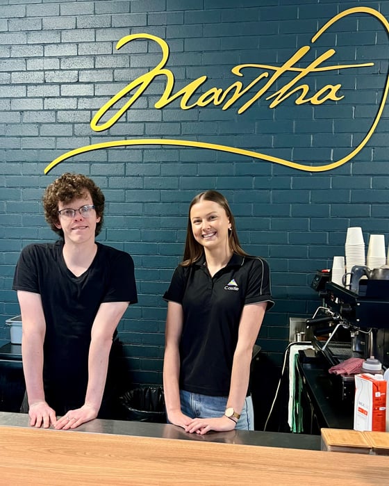 Lochlan and Amy are smiling behind a counter next to a coffee machine. Lochlan is wearing a black tshiirt, glasses and has brown curly hair.