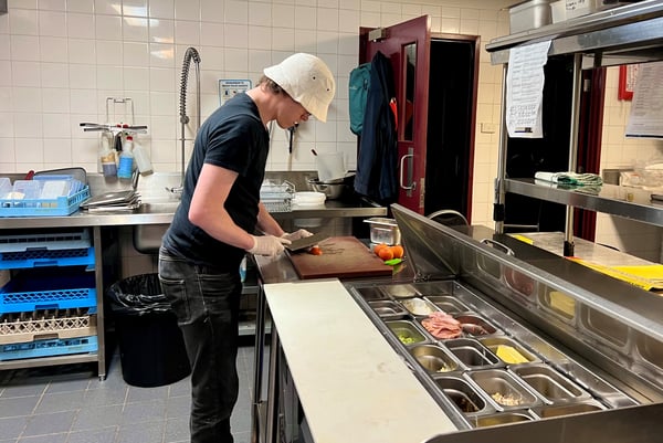 Lochlan is standing in the kitchen at a food prep bench. He is chopping vegetables with a large knife. He is wearing black pants, black t-shirt, a white hat and food safe gloves.
