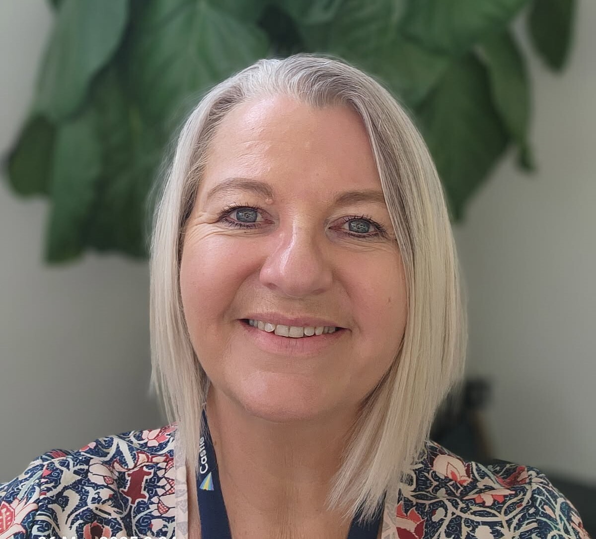 Headshot of Schon, a woman in her 50s with short blonde hair smiling at the camera. She wears a floral top with a Castle lanyard.