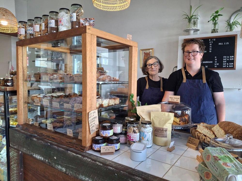 [Picture of Louise and Hannah smiling at the counter of Burnt Honey Bakery in Long Jetty, with a cabinet of pastries to the left, and jam jars, fresh bread and eggs on the counter. Louise and Hannah are wearing navy blue aprons.]