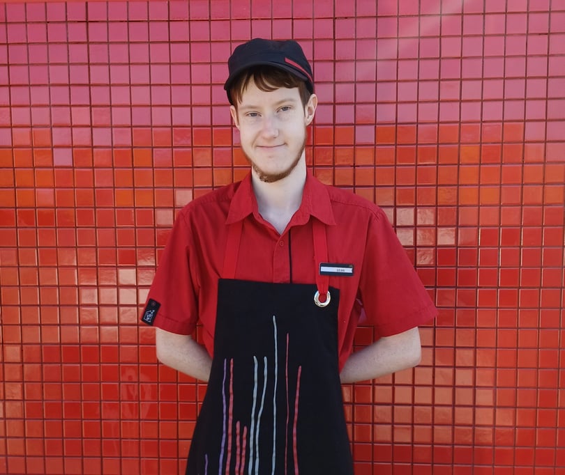 Sean, a young man wearing his McDonald's uniform of red shirt and black apron and cap, standing in front of red tiled background