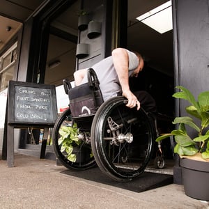 A man is pushing a wheelchair up a small rubber ramp into a cafe doorway.